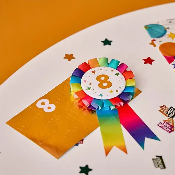 Colourful number 8 birthday badge with rainbow ribbons displayed on a party table surrounded by confetti and birthday decorations.